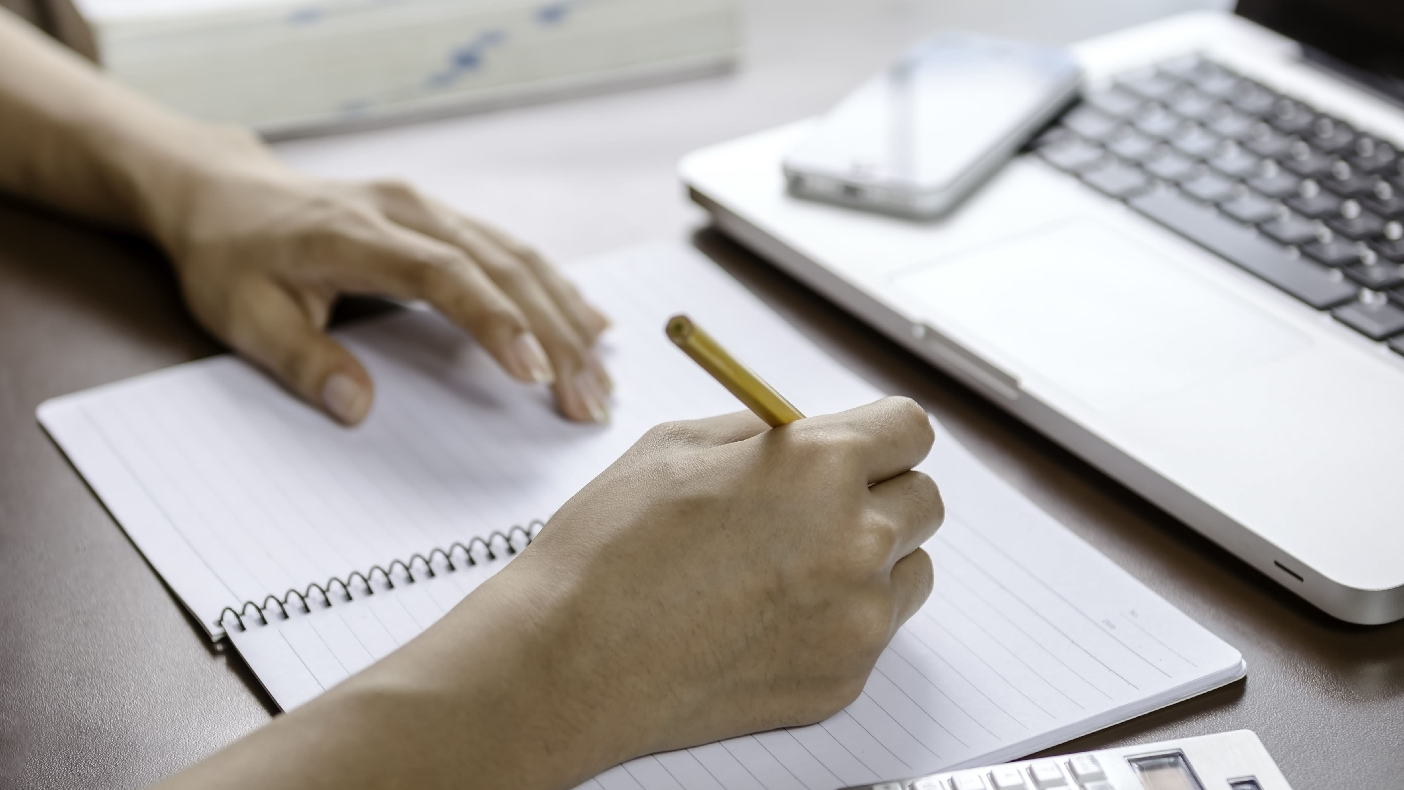 Person writing in notepad with laptop and phone out