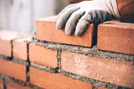Construction worker placing a red brick on a wall with mortar, symbolizing building, labor, or foundation work.