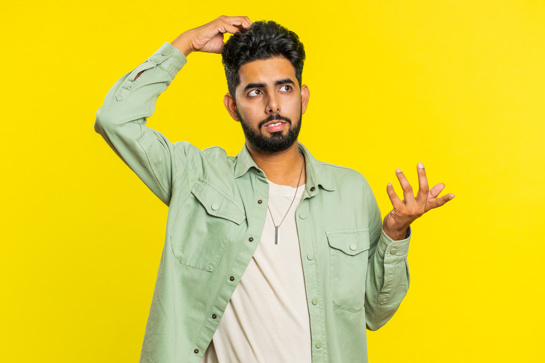Confused man scratching his head and shrugging, standing against a bright yellow background, looking uncertain.